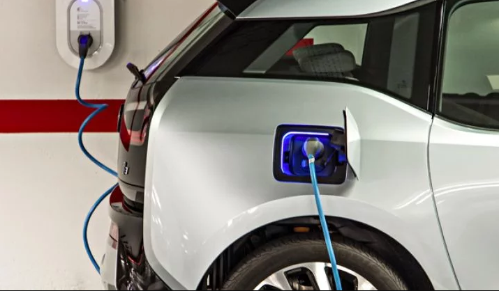 This image shows a white electric vehicle being charged at a charging station, with a blue charging cable connected from the wall-mounted charger to the car's charging port.