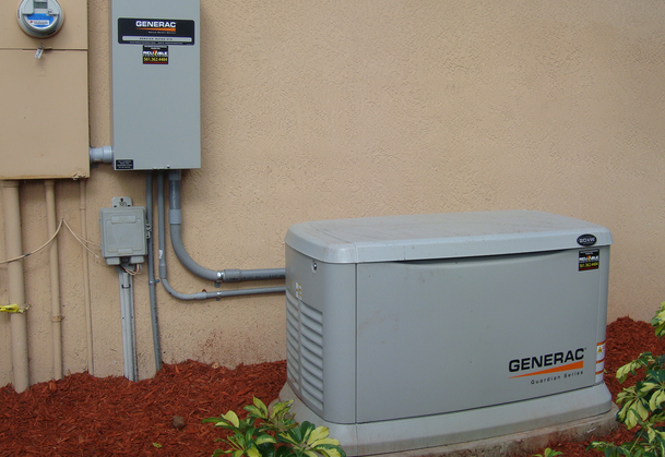 This image shows a Generac backup generator system installed outside a home, with the main generator unit on the ground connected to a transfer switch mounted on the exterior wall.
