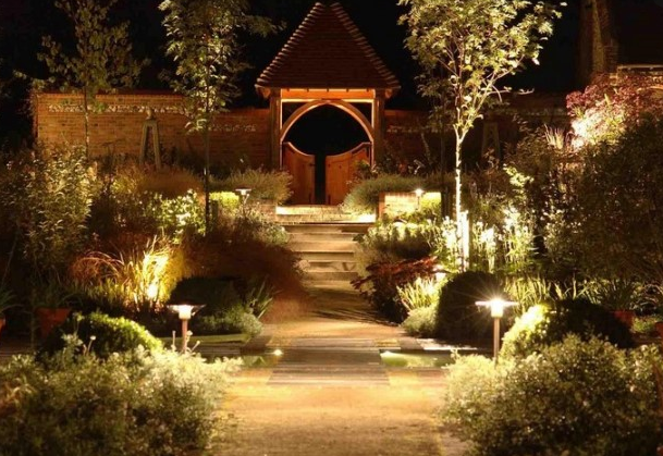 This image shows a beautifully illuminated garden pathway at night leading to a wooden pavilion or gazebo, with strategically placed landscape lighting highlighting the walkway, plants, and architectural features.