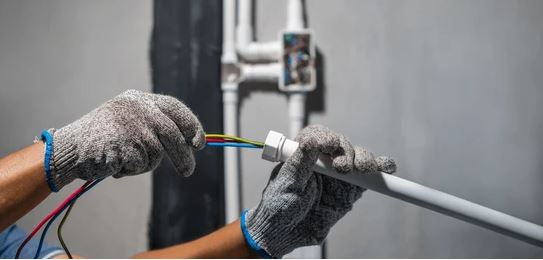 This image shows a person's hands wearing protective gloves working with electrical wiring, stripping or connecting colored wires to what appears to be electrical conduit or cable.