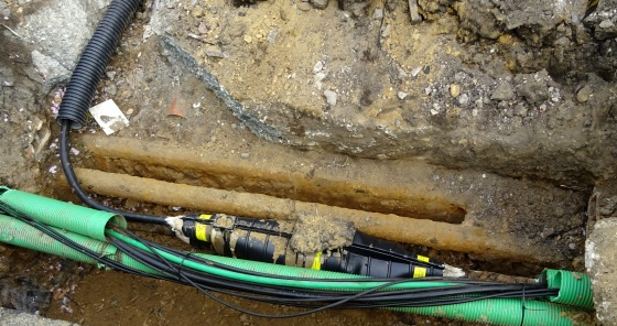 This image shows underground utility work with green conduit pipes and electrical cables with splice connections laid in an excavated trench alongside what appears to be an existing clay pipe.