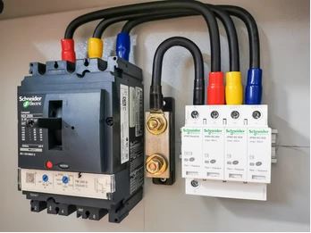 This image shows electrical components including a black circuit breaker or contactor on the left connected by cables to white electrical devices (likely surge protectors or disconnect switches) mounted on a panel.