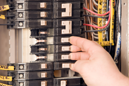 This image shows a person's hand reaching to operate or adjust a circuit breaker in an electrical panel filled with multiple breakers and colorful wiring.