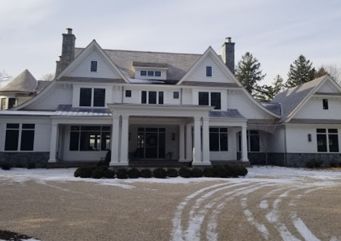 This image shows a large, modern white house with dark trim and multiple gables, featuring a covered front entrance with columns and surrounded by snow-covered ground and evergreen trees.